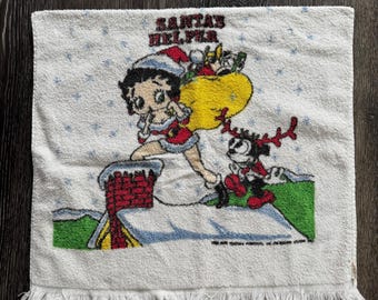 Op de afbeelding: Witte kersthanddoek met Betty Boop als Santa's helper. De handdoek heeft een gefranjerde rand en een herhalend blauw sterrenpatroon. Het ontwerp omvat een schoorsteen, een rendier en een gele zak met cadeaus.