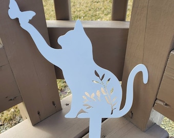 May include: White metal silhouette of a cat reaching up to catch a butterfly. The cat has a floral design on its side and a long tail. The silhouette is attached to a metal stake.