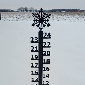 May include: A black metal snowflake snow gauge with a numbered scale from 2 to 24. The gauge is standing in a snowy field.