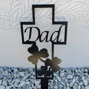 May include: Black metal cross with the word "Dad" and shamrock designs. The cross is on a bed of gray gravel.