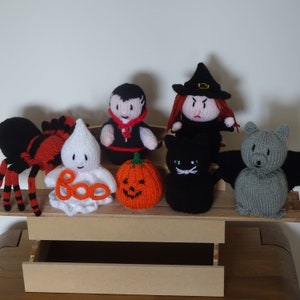 May include: A selection of handmade knitted Halloween characters. The collection features a black spider, a white ghost with "boo" text, a vampire, a witch, a black cat, an orange pumpkin, and a grey bat. Each doll has distinct features.