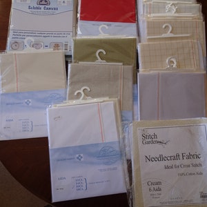 May include: Selection of cross-stitch fabrics in different colours and sizes, including white, red, and cream. Some fabrics are packaged with hangers. The image also shows soluble canvas and Aida cloth, ideal for needlecraft projects.