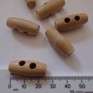May include: Several light brown wooden toggle buttons, each with two holes. The buttons are cylindrical with rounded ends. A ruler with millimetre markings is in the foreground.