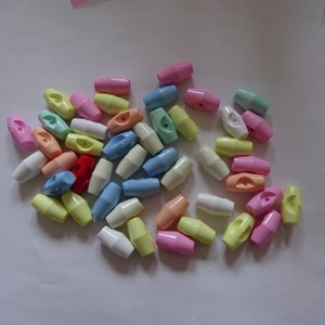 May include: A selection of pastel-coloured plastic cord locks. The oval-shaped fasteners are in shades of pink, yellow, blue, and white. Each has a central hole for cord insertion. These are small craft components.