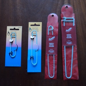 May include: Four safety stitch holders in varying sizes. Two are packaged on blue cards with the text "Pony" and "Stitch Holder". The other two are in red packaging with the text "Essentials Safety Stitch Holder". All are light blue.