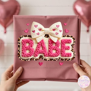 Puede incluir: Un artículo de tela rosa con un gran aplique "BABE" texturizado en rosa brillante, delineado con un borde de estampado de leopardo. Un lazo color crema con detalles de corazones rosas está encima de la palabra. Pequeños corazones rosas están dispersos.