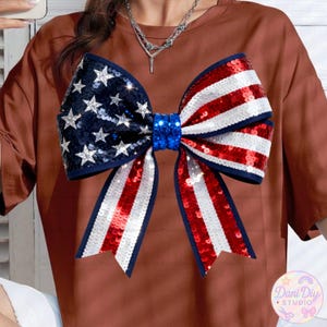 Peut inclure: Un t-shirt marron orn&eacute; d'un grand n&oelig;ud orn&eacute; de paillettes aux couleurs patriotiques. Le n&oelig;ud reprend le drapeau am&eacute;ricain avec des paillettes bleues, blanches et rouges. Le n&oelig;ud est centr&eacute; sur le t-shirt, cr&eacute;ant un look festif et accrocheur.