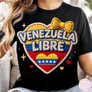 Puede incluir: Camiseta negra con un diseño en forma de corazón con las palabras "VENEZUELA LIBRE". El corazón incorpora los colores de la bandera venezolana, con un lazo amarillo en la parte superior. Pequeños acentos de estrellas y corazones adornan el diseño.