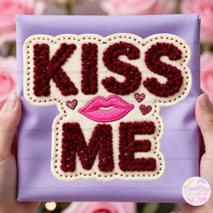Puede incluir: Una caja de regalo lavanda con una aplicación decorativa que dice "KISS ME". La aplicación presenta texto burdeos, un diseño de labios rosas y pequeños acentos de corazón. La aplicación está delineada con un borde color crema.