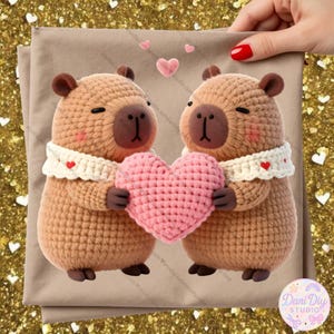 Puede incluir: Dos carpinchos de ganchillo sosteniendo un corazón rosa. Llevan bufandas blancas con corazones rojos. Los carpinchos son de color marrón claro con orejas y narices marrones oscuras. El fondo es una tela beige con pequeños corazones rosas.