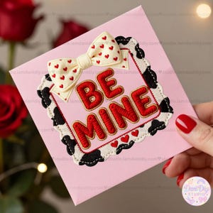 Puede incluir: Una tarjeta rosa con las palabras bordadas "BE MINE" en rojo, enmarcada por un borde con estampado de vaca en blanco y negro. Un lazo crema con corazones rojos adorna la parte superior. La tarjeta es sostenida por una mano con esmalte de uñas rojo.