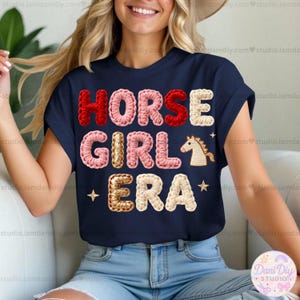 Puede incluir: Camiseta azul marino con el texto "HORSE GIRL ERA" en estilo crochet. El texto es rojo, rosa, dorado y crema, con un gráfico de caballo y estrellas. Una camiseta informal con temática ecuestre.