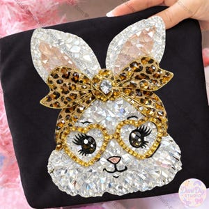 Puede incluir: Tela negra con un diseño de conejo brillante. El conejo tiene detalles de cristal, un lazo con estampado de leopardo y gafas en forma de corazón con monturas doradas. El diseño incluye una pequeña nariz rosa y ojos negros.