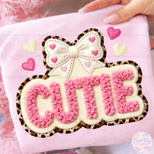 Puede incluir: Una camiseta rosa claro con un parche bordado grande que dice "CUTIE" en letras rosas texturizadas. El parche tiene un borde con estampado de leopardo y un lazo con detalles de corazón. Apliques de corazón adicionales están dispersos.