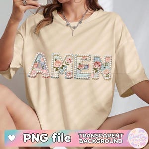 Puede incluir: Una camiseta color crema con la palabra "AMEN" bordada en la parte delantera. Las letras están decoradas con motivos florales y delineadas con perlas de imitación. La camiseta también tiene el texto "PNG file" y "TRANSPARENT BACKGROUND".