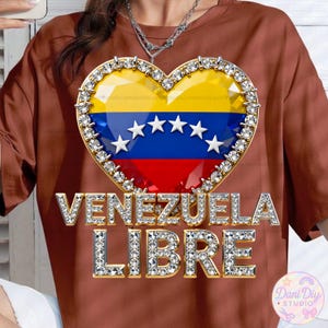May include: A brown t-shirt featuring a heart-shaped design with the Venezuelan flag and the words "VENEZUELA LIBRE" in a sparkling, diamond-like font. The heart is outlined with a gold border and embellished with rhinestones.