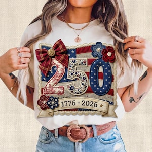 Puede incluir: Camiseta color crema con un diseño patriótico con el número "250" en rojo, blanco y azul. El diseño incluye detalles florales, un lazo y el texto "1776-2026" en una pancarta. La camiseta se combina con vaqueros azules y un cinturón marrón.