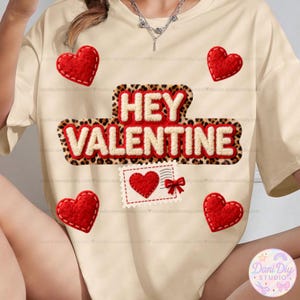 Puede incluir: Camiseta color crema con temática de San Valentín. La camiseta presenta el texto "HEY VALENTINE" en un diseño decorativo, junto con varias aplicaciones de corazón rojo y un gráfico de sello postal con un corazón y un lazo.