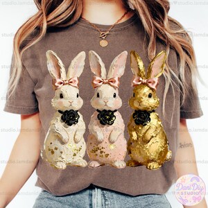 Puede incluir: Tres figuras decorativas de conejitos de Pascua. Una es blanca con detalles dorados, otra rosa y la tercera dorada. Cada conejito tiene un lazo rosa y un collar de rosa negra. Una encantadora decoración de Pascua.