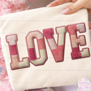 May include: Cream-coloured sweatshirt featuring the word "LOVE" in a patchwork design. The letters are in shades of pink, cream, and peach with a fuzzy texture and a brown border. A hand holds the sweatshirt.