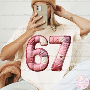 May include: A cream-colored t-shirt featuring the number "67" in a patchwork design. The numbers are in shades of pink and white, with heart accents on the "7". The image is of a person holding a phone.