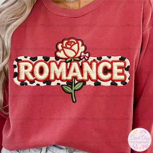 Puede incluir: Una sudadera roja con la palabra "ROMANCE" bordada en blanco con un borde moteado en blanco y negro. Una rosa color crema con un lazo rosa y un tallo verde está encima de la palabra.