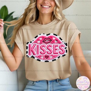 May include: A tan t-shirt featuring a large, embroidered design of pink lips with the word "KISSES" in pink lettering. The design includes small hearts and a black and white border. The shirt is a casual style.