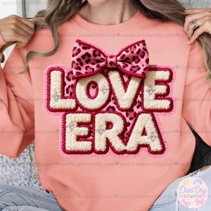 Puede incluir: Sudadera rosa claro con las palabras "LOVE ERA" en una fuente blanca texturizada con contorno rojo. Un lazo rosa con estampado de leopardo está encima del texto, con pequeños diamantes de imitación en el diseño.