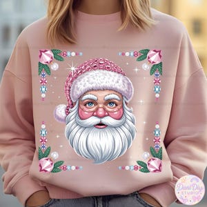 Puede incluir: Sudadera rosa claro con un diseño de Papá Noel. El Papá Noel lleva un gorro y gafas rosas, y tiene barba blanca. El diseño está enmarcado con gemas rosas y hojas verdes, ideal para la temporada navideña.