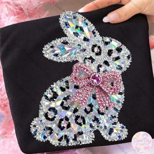 Puede incluir: Tela negra con un diseño de conejo de pedrería con estampado de leopardo. El conejo tiene un lazo rosa con una joya central y está adornado con pedrería iridiscente.