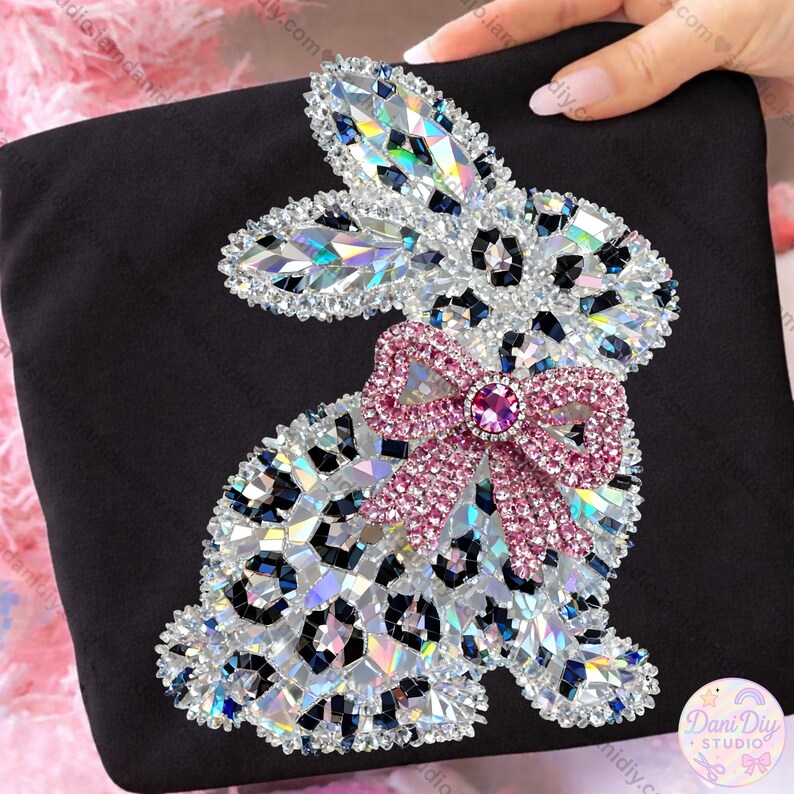 May include: Black fabric featuring a rhinestone bunny design with a leopard print pattern. The bunny has a pink bow with a central jewel. The design is embellished with iridescent rhinestones.