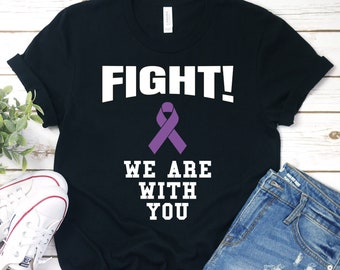 May include: Black t-shirt with white text that reads "FIGHT! We are with you". A purple ribbon is above the text. The shirt is laying on a wooden surface with a pair of white sneakers and blue denim shorts.