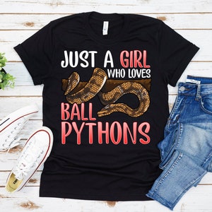 May include: Black t-shirt with the text "Just a girl who loves ball pythons" in red and white. A brown and white ball python is pictured on the shirt.