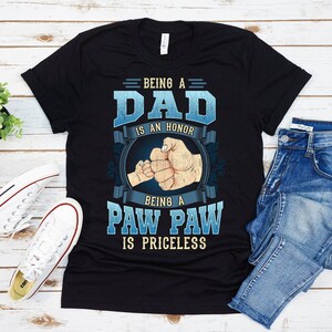 May include: Black t-shirt with a blue and white graphic design. The graphic features two fists bumping, with the text "Being a Dad is an Honor, Being a Paw Paw is Priceless".