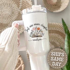 May include: A white stainless steel tumbler with a handle and a clear lid. The front features a graphic of an open book with colorful flowers emerging from the pages, the text "Just one more chapter", and the personalized name "madison" below.