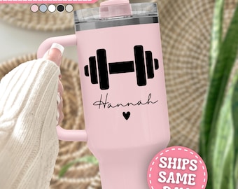 Puede incluir: Un vaso de acero inoxidable de color rosa claro con asa, tapa transparente y pajita. El vaso presenta un gráfico negro de una mancuerna, el nombre Hannah en letra cursiva negra y un pequeño icono de corazón negro bajo el nombre.