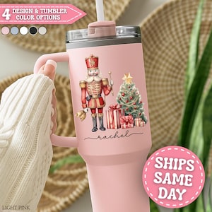 May include: A light pink tumbler with a handle, featuring a Christmas design. The design includes a nutcracker, Christmas tree, and wrapped gifts. The text "Rachel" is written in cursive. The top of the tumbler has a straw and a lid. The text "4 DESIGN & TUMBLER COLOR OPTIONS" is visible.