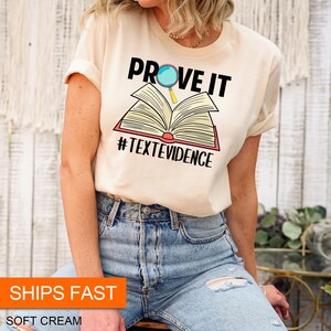 Puede incluir: Una camiseta de color crema con el texto "PROVE IT" y una lupa sobre un libro abierto. El texto "#TEXTEVIDENCE" est&aacute; debajo del libro.