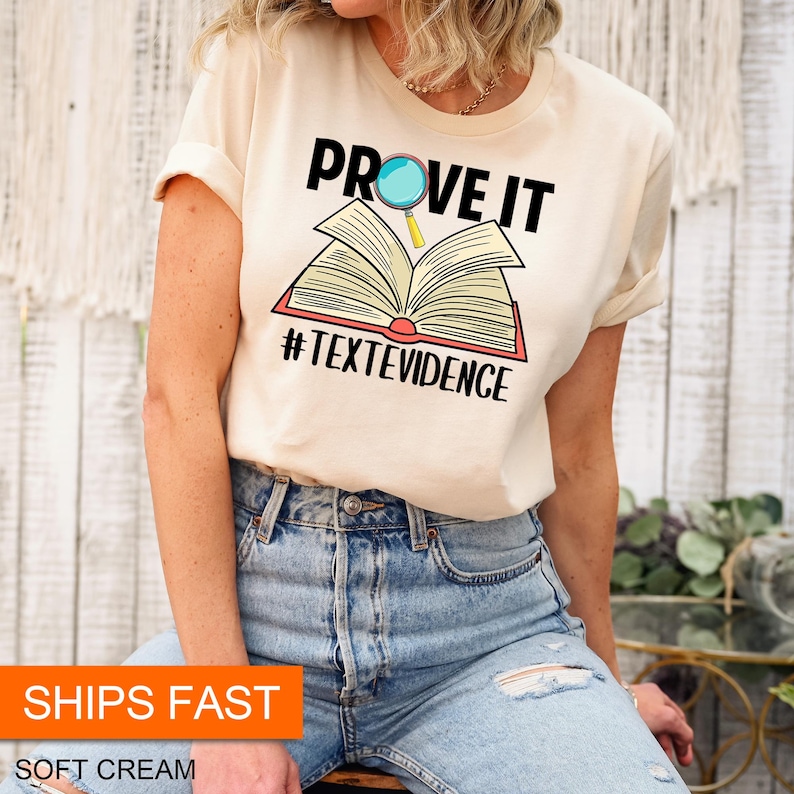 Puede incluir: Una camiseta de color crema con el texto "PROVE IT" y una lupa sobre un libro abierto. El texto "#TEXTEVIDENCE" est&aacute; debajo del libro.