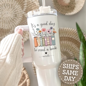 May include: A white stainless steel tumbler with a handle and a clear lid. The design features a row of colorful books with flowers growing from the top, the text "It's a good day to read a book," and the name "michelle" in cursive script.