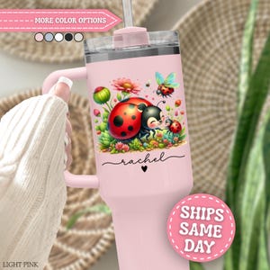May include: A light pink tumbler with a handle and a clear lid featuring a graphic of a large ladybug and a smaller ladybug among colorful flowers and greenery. The name rachel is printed in black script below the illustration with a small heart.