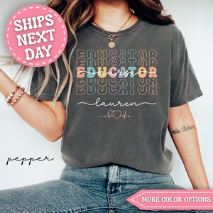 May include: Dark gray t-shirt with the words "Educator Nurse" in colorful, stacked text. The name "Lauren" is written below in cursive, with a nurse symbol and heartbeat graphic. A pink circle with "SHIPS NEXT DAY" is in the upper left.