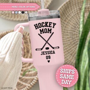 May include: A light pink stainless steel tumbler with a handle and a clear lid. The front features black text reading HOCKEY MOM, JESSICA, and 09, along with a graphic of two crossed hockey sticks, a puck, and a small heart symbol below the number.