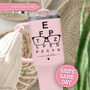 May include: A light pink insulated tumbler with a handle and a clear lid featuring a black eye chart graphic with glasses, the name michelle in script, and a small heart icon. The tumbler has a straw inserted through the top lid opening.