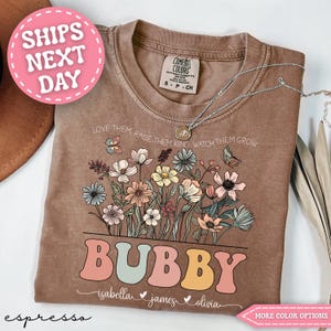 May include: Espresso-colored t-shirt with a floral design and the word "BUBBY" in pastel colors. The shirt features the text "LOVE THEM, RAISE THEM KIND, WATCH THEM GROW" and names below. A pink circle with "SHIPS NEXT DAY" is visible.