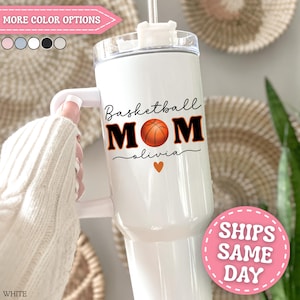 May include: White tumbler with a handle, featuring the text "Basketball MOM" in black and orange, with a basketball graphic. Below the text is the name "olivia" and a small orange heart. The tumbler has a clear lid and a straw.