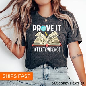 Puede incluir: Camiseta de color gris oscuro jaspeado con el texto "PROVE IT" y un libro abierto con una lupa sobre &eacute;l. El texto "#TEXTEVIDENCE" est&aacute; debajo del libro.