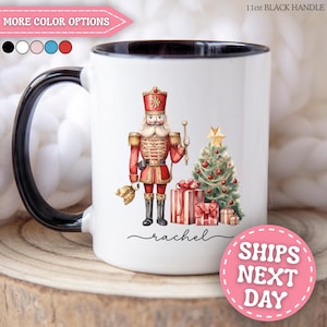Puede incluir: Una taza de cerámica blanca de 312 ml con asa negra que presenta un gráfico de un cascanueces con uniforme rojo junto a un árbol de Navidad decorado y regalos. El nombre rachel está impreso en letra cursiva negra bajo la ilustración.