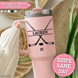 May include: A light pink tumbler with a handle, decorated with a hockey theme. The design includes crossed hockey sticks, a puck, and the name "LAUREN" above the number "21". The tumbler has a straw and a lid. The text "MORE DESIGN & TUMBLER COLOR OPTIONS" is visible.