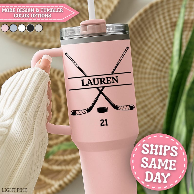 May include: A light pink tumbler with a handle, decorated with a hockey theme. The design includes crossed hockey sticks, a puck, and the name "LAUREN" above the number "21". The tumbler has a straw and a lid. The text "MORE DESIGN & TUMBLER COLOR OPTIONS" is visible.